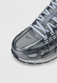 Αθλητικό παπούτσι σε ασημί χρώμα με πλέγμα στις επιφάνειες και λαμπερές λεπτομέρειες. Διαθέτει λευκά κορδόνια και μαύρη καουτσούκ σόλα. Εμφανές διάτρητο μοτίβο στο πάνω μέρος.