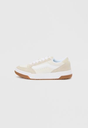 Alacsony szárú, bézs és fehér sportcipő fehér fűzőkkel és gumitalpakkal, külső oldalról, egyszínű háttéren.