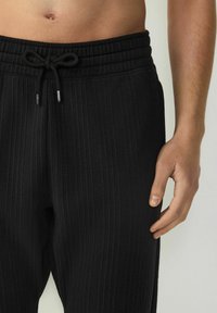 Los pantalones de chándal negros con canalé presentan una cinturilla elástica con cordón. El tejido parece suave con una textura lisa.