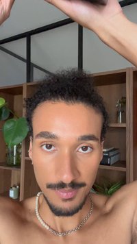 Gros plan d'une personne aux cheveux bouclés, portant un collier de perles et une boucle d'oreille, devant un fond en bois simple avec des plantes.