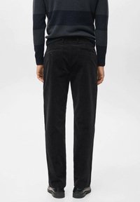 Pantaloni di velluto a coste neri con un design a gamba dritta, tasche posteriori con dettagli verdi, indossati con un maglione a righe blu navy e scarpe nere.
