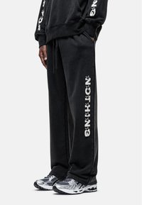 Zwarte sweatpants van een katoenmix, voorzien van een elastische tailleband met trekkoord en gedurfde witte grafische tekst op het linkerbeen. Gedeeltelijk met metallic sneakers.