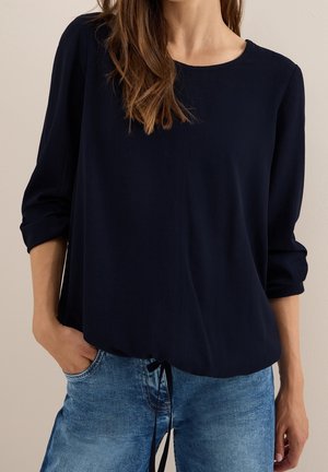 Kvinde iført mørkeblå bluse med lange ærmer med opkrogede ærmer og blå denimjeans, hånd i lommen, mod neutral baggrund.