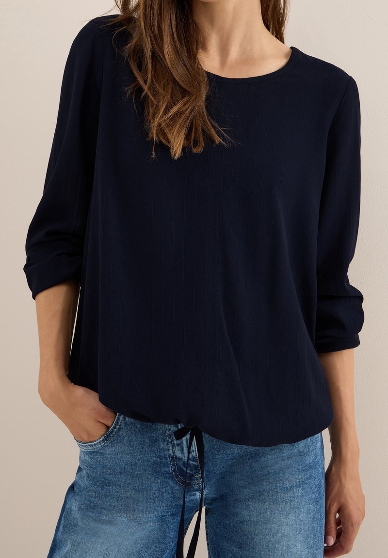 Femme portant un chemisier bleu foncé à manches longues retroussées et un jean bleu en denim, main dans la poche, sur fond neutre.