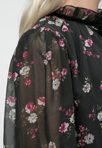 Blusa nera trasparente con maniche, motivo floreale rosa e bianco, colletto a volant e dettagli sulle spalle a palloncino.