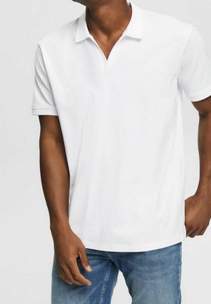 Mand iført en ensfarvet hvid kortærmet poloshirt og blå jeans, stående med den ene hånd i lommen mod en hvid baggrund.