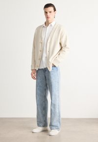 Beige pletený cardigan se třemi knoflíky, zkombinovaný s bílou košilí a světle modrými džíny. Sadu doplňují jednoduché bílé tenisky.