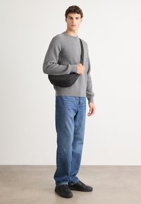 Gray ribbed sweater, blue denim jeans, black shoulder bag, and black sneakers. Model stands on a smooth, neutral-toned floor with plain background.