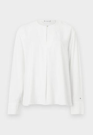 Camisa branca de mangas longas com decote redondo, detalhe de pregas frontais e fecho de botão, com mangas com punhos e textura de tecido suave.