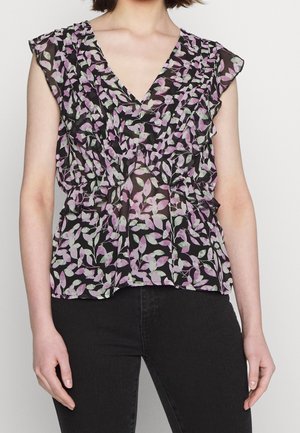 Frau trägt ärmellose schwarze Bluse mit lila-weißem Blattmuster und mehrlagigen Rüschen-Details, kombiniert mit schwarzen Hosen.