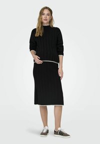 Pull en maille côtelée noir à manches 3/4, avec un biais blanc au cou et à l'ourlet, associé à une jupe côtelée noire assortie et des chaussures sportives.