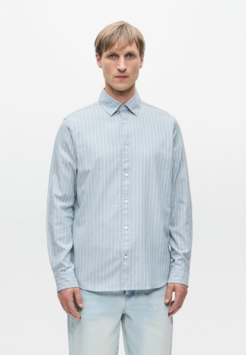 Ανοιχτό μπλε πουκάμισο με μακριά μανίκια και λεπτές κάθετες λευκές ρίγες, με κουμπωτό γιακά και μπροστινή κουμπωτή κλείσιμο, σε συνδυασμό με ανοιχτό μπλε τζιν.