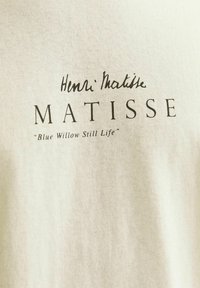 Hellbeige Baumwoll-T-Shirt mit aufgedrucktem Text: "Henri Matisse" über "MATISSE" und "Blue Willow Still Life" in einer schlichten schwarzen Schriftart.