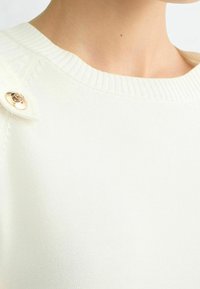 Světlounce krémový pletený svetr s výrazným vzorem, s žebrovaným výstřihem a zlatým knoflíkem poblíž ramene.