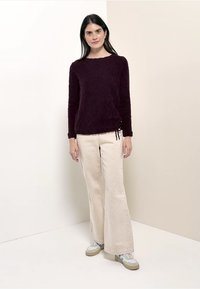 Fioletowy sweter o fakturowanej strukturze z okrągłym dekoltem, zestawiony z kremowymi spodniami o szerokich nogawkach oraz szarymi sneakersami. Sweter posiada regulowane wiązania po bokach.