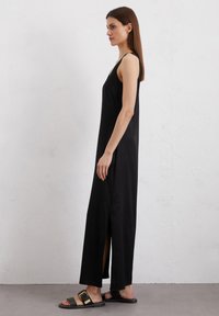 Robe maxi sans manches noire avec une fente sur le côté, fabriquée en tissu lisse. Portée avec des sandales plates noires avec un détail de boucle dorée.