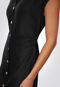 Vestido negro de botones con mangas cortas, que presenta un detalle anudado en la cintura y acentos de botones de madera en la parte delantera. Textura suave.