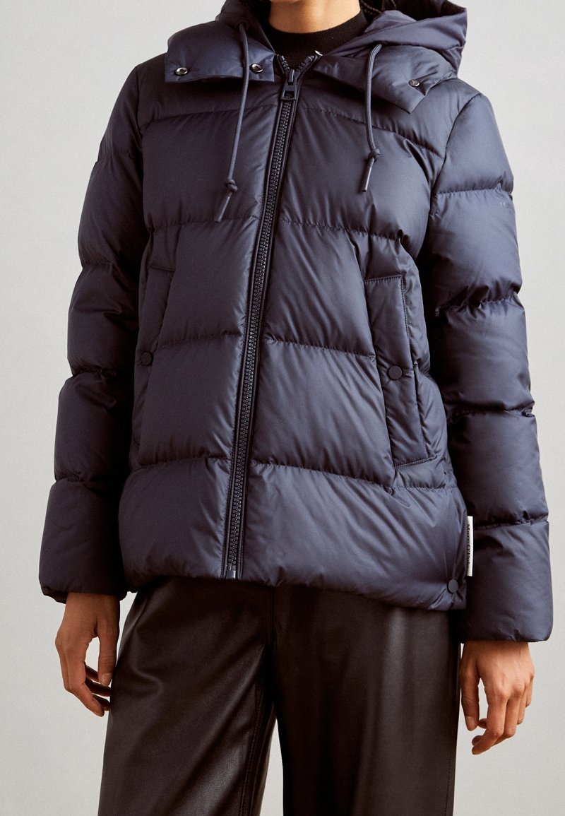 Personne portant une veste matelassée bleu marine avec fermeture éclair à l'avant, poches à boutons-pression et capuche avec cordons, associée à un pantalon foncé.