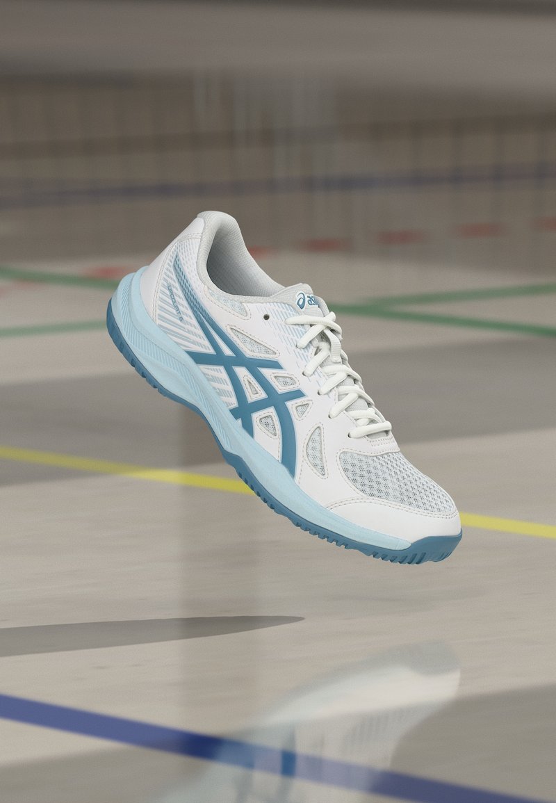 Άσπρο και γαλάζιο αθλητικό παπούτσι με κορδόνια, αιωρούμενο πάνω από το δάπεδο ενός εσωτερικού αθλητικού γηπέδου.