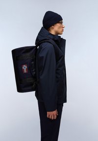 Námořnická modrá voděodolná bunda, tmavá čepice a tmavé kalhoty. Na sobě má černý batoh s výrazným logem a detaily vlajky. Pohled z boku.