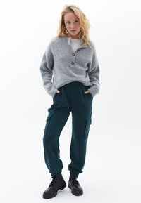Cardigan grigio a maglia con tre bottoni, abbinato a pantaloni cargo tapered verde scuro con tasche laterali, e stivali neri con lacci.