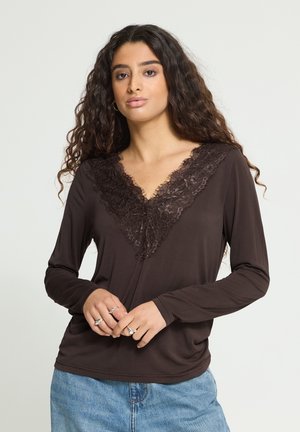 Femme aux longs cheveux bouclés portant un haut marron à manches longues avec un décolleté en V en dentelle et un jean bleu, tenant ses mains jointes devant elle.