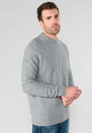 Pull gris à côtes avec manches longues et col rond, présentant un motif de tricot texturé et des poignets ajustés. porté sur un jean bleu.
