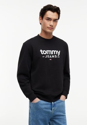 Młody mężczyzna w czarnej bluzie Tommy Jeans z logo, jasnoniebieskie dżinsy, stojący z rękami w kieszeniach na tle jednolitego, białego tła.
