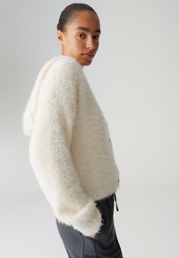 Weiße, flauschige Strickkapuzenjacke mit lockerem Schnitt, angesetzten Schultern und gerippten Bündchen. Trägt man über dunkler Hose, vor einfarbigem Hintergrund.