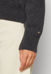 Maglione lavorato a maglia grigio scuro con polsini a costine, caratterizzato da un piccolo emblema con accenti rossi e bianchi sulla manica. Pantaloni chiari sottostanti.