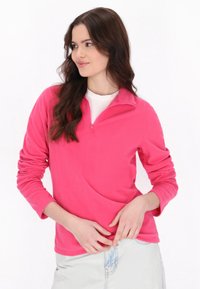 Pullover in pile, di colore rosa brillante, con mezza zip sul collo, maniche lunghe, dalla texture leggera, indossato con una camicia bianca e jeans in denim chiaro.