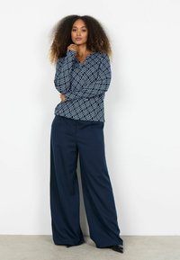 Tmavomodrá blúzka s dlhým rukávom a geometrickým bielym vzorom, skombinovaná so širokými tmavomodrými nohavicami. Hladká látka a pohodlný strih.