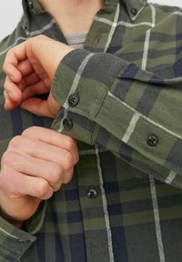 Grün-kariertes Flanellhemd in Marineblau mit Knöpfen an den Bündchen. Es verfügt über eine weiche Textur und ein klassisches Kragendesign, dargestellt durch eine Hand, die den Ärmel anpasst.