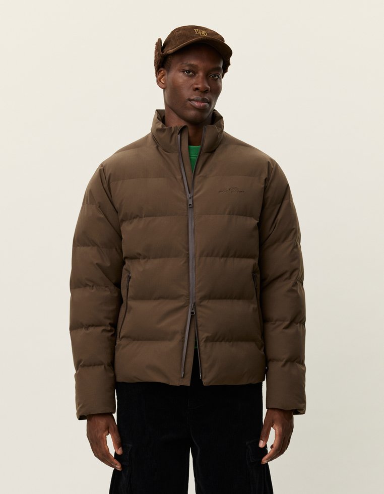 Brun puffer-jakke med glidelås, med høy krage og quiltet design. Utstyrt med sidelommer og laget av glatt stoff.