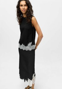 Robe noire sans manches avec bordure en dentelle à l'ourlet, présentant un tissu texturé et une silhouette ajustée. La dentelle est blanche avec un bord festonné.