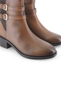 Botas de cuero marrón hasta el tobillo con punta afilada, que presentan acentos de doble hebilla y paneles laterales elásticos para un fácil uso. Tacón plano negro.