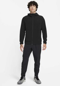 Zwarte jas met ritssluiting en capuchon, gemaakt van lichtgewicht stof. Gecombineerd met zwart nauwsluitende broek en sportieve schoenen met zilverkleurige accenten.