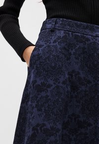 Μπλε σκούρο φούστα με περίπλοκο σκούρο λουλουδάτο σχέδιο, υφή υφάσματος και προσαρμοσμένη μέση. Είναι ορατή μια λεπτομέρεια πλαϊνής τσέπης.
