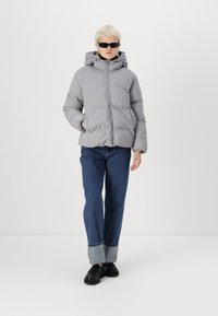 Grå vadderad jacka med huva, med en framsida med dragkedja och två sidofickor. Bärs med vida blå jeans och svarta skor.