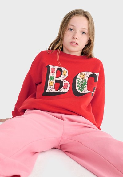 Ragazza giovane con capelli biondi indossa una felpa rossa con lettere floreali colorate "BC" e pantaloni rosa, seduta contro uno sfondo chiaro e semplice.