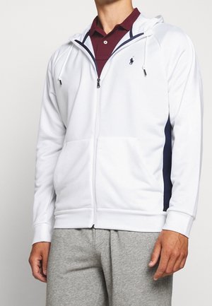 Man draagt een wit hoodie met ritssluiting en marineblauwe zijpanelen over een bordeauxrode polo en grijze trainingsbroek, staand tegen een effen achtergrond.