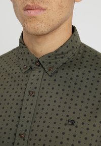 Oliwkowa koszula zapinana na guziki z czarnymi okrągłymi wzorami, brązowymi guzikami i subtelnym logo. Posiada klasyczny kołnierz i teksturowaną tkaninę.