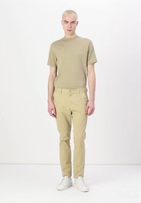 Camiseta de algodón beige de mangas cortas, combinada con pantalones slim-fit beige claros y zapatillas blancas, de pie frente a un fondo liso.