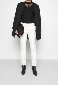 Svart åtsittande topp med nätinslag, lager under en svart trenchcoat. Sma vita byxor och svarta spetsiga ankelskor, håller en svart väska.