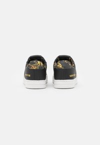 Pair of black low-top sneakers with white soles, gold baroque pattern on heel and collar, and "VERSACE JEANS COUTURE" logo in gold.