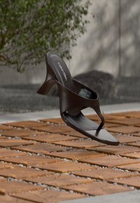 Μαύρο πέδιλο Calvin Klein με τακούνι, τετράγωνη μύτη και λεπτό λουράκι που αιωρείται πάνω από επιφάνεια εξωτερικού χώρου από τούβλα και πέτρες.