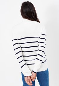 Donna con lunghi capelli scuri di spalle, che indossa un maglione bianco con righe orizzontali nere e jeans blu, con le mani unite dietro la schiena.