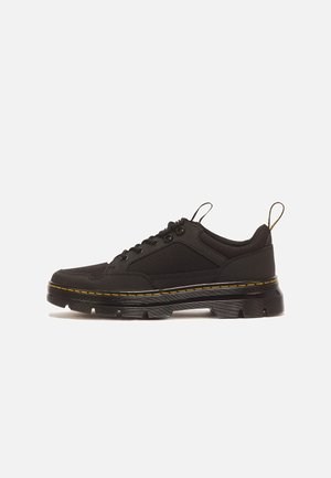 Chaussure basse noire à lacets avec semelle épaisse, surpiqûres jaunes et languettes au talon et à la languette sur fond blanc.