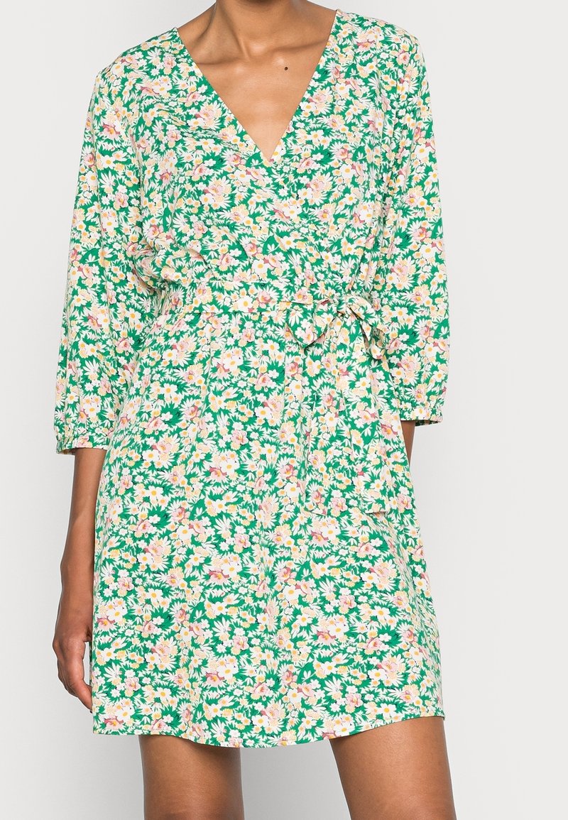 Femme portant une robe portefeuille verte à fleurs roses et blanches, avec des manches trois quarts et une ceinture nouée à la taille.