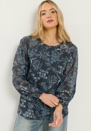 Kvinde justerer ærmet på navyblå bluse med lyseblåt blomsterprint og gennemsigtige pufærmer, iført lyseblå jeans, stående mod en hvid væg.
